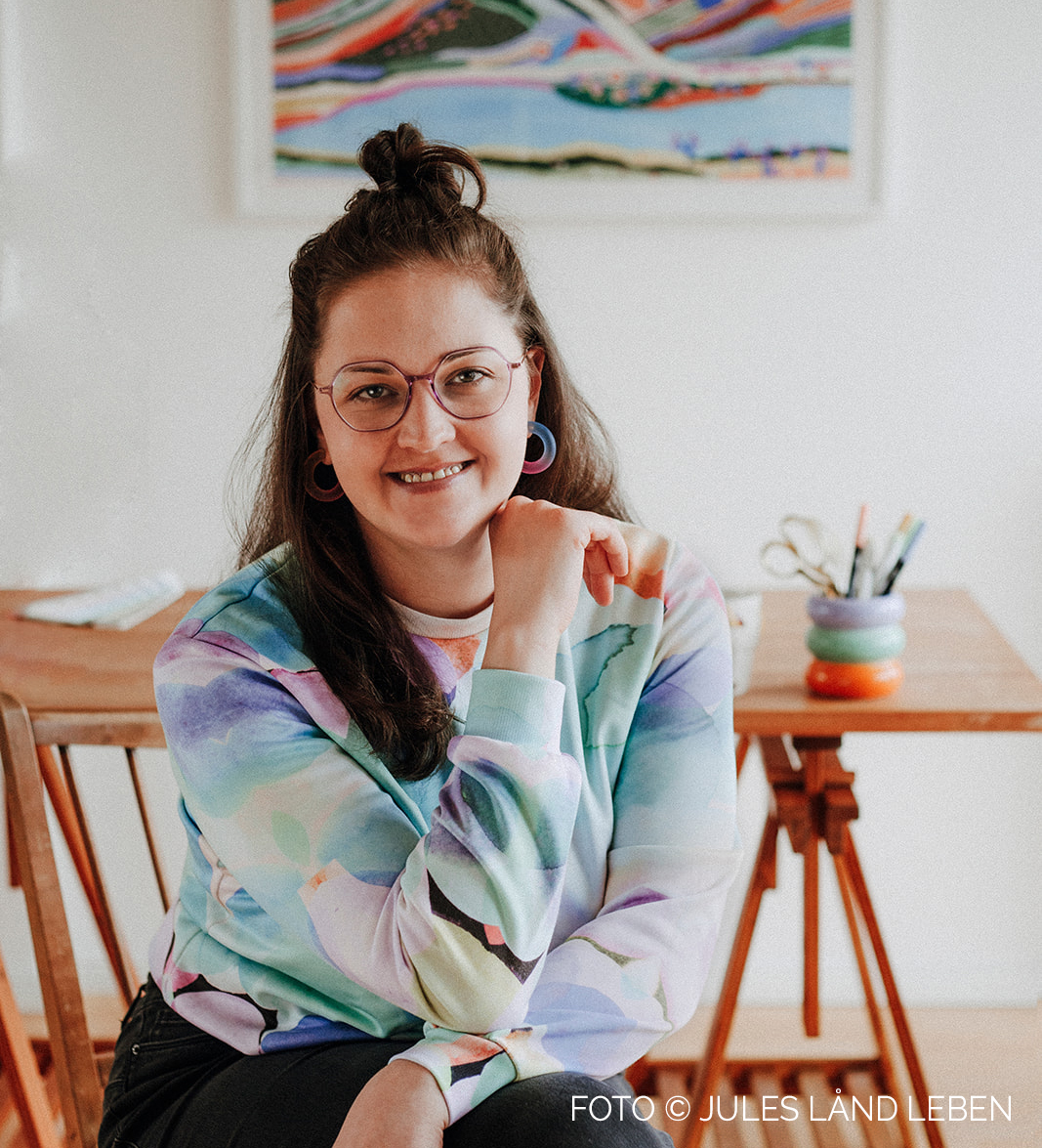 Profilfoto von Eva, wie sie fröhlich strahlend auf ihr Knie gestützt vor ihrem Schreibtisch sitzt.