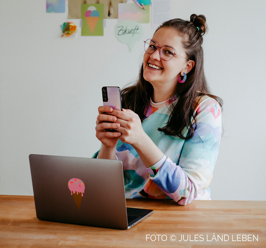 Grafikdesignerin Eva Lisette Zahneißen sitzt vor ihrem Laptop mit einem Smartophone in der Hand und lacht freundlich in die Kamera.